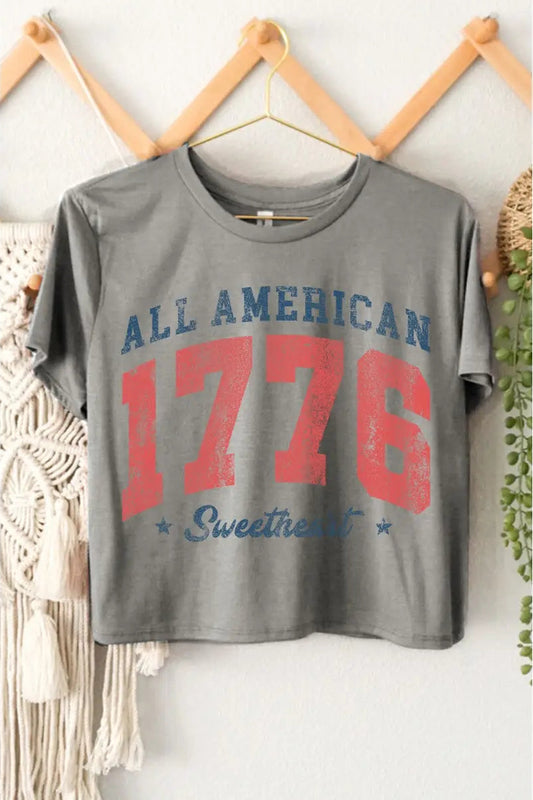 Gray t-shirt with 'All American 1776 Sweetheart' text on a hanger against a white wall.