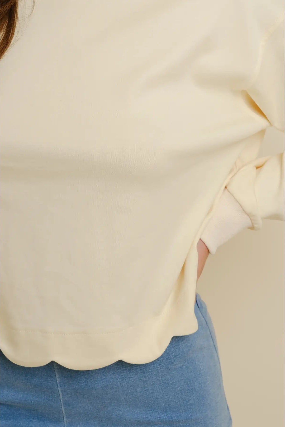 Close-up of a person wearing a cream-colored long-sleeve top with a scalloped hem and blue jeans against a plain beige background.