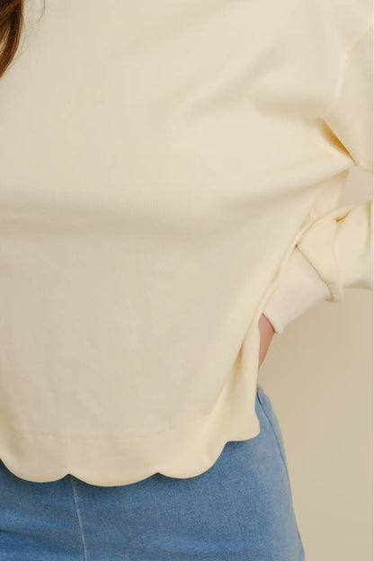 Close-up of a person wearing a cream-colored long-sleeve top with a scalloped hem and blue jeans against a plain beige background.