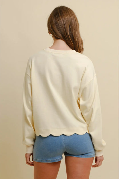 A woman viewed from the back wearing a cream-colored long-sleeve crewneck sweatshirt with a scalloped hem and light blue denim shorts, set against a solid beige background.