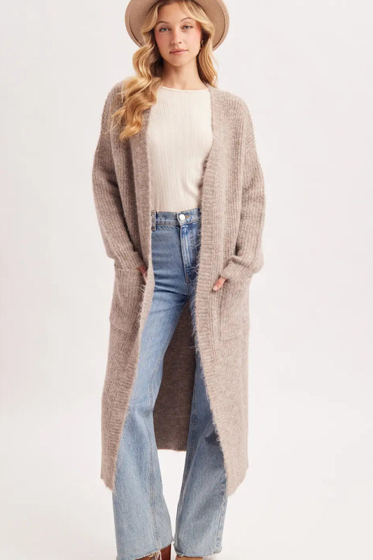 A woman modeling a long, open-front, oversized knit cardigan in a light taupe-beige color, styled with a cream top, blue jeans, and a beige hat against a plain light background.