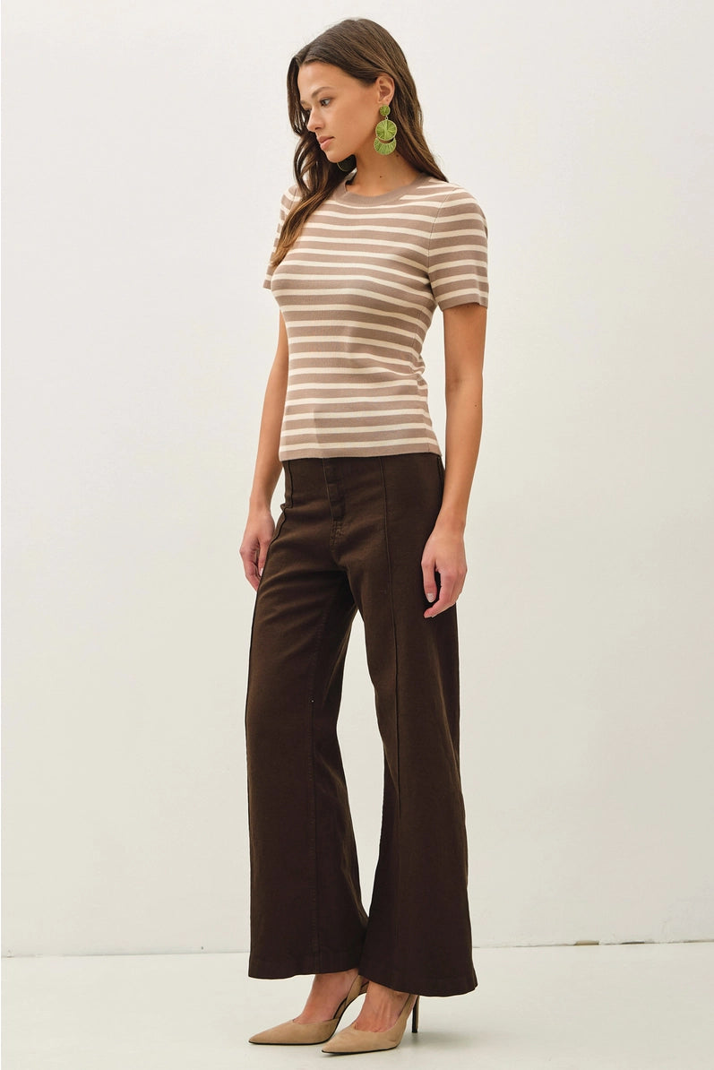 A woman modeling a short-sleeve crewneck top with beige and brown horizontal stripes, paired with dark brown wide-leg pants and beige heels, against a plain white studio background.