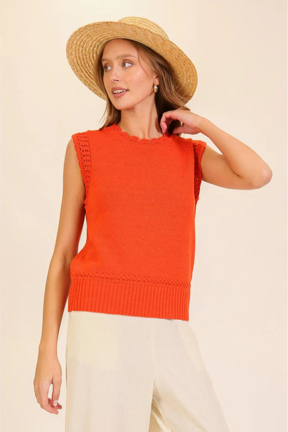 A woman wearing a sleeveless bright orange knit vest, cream wide-leg pants, and a straw hat, posing against a solid beige background.