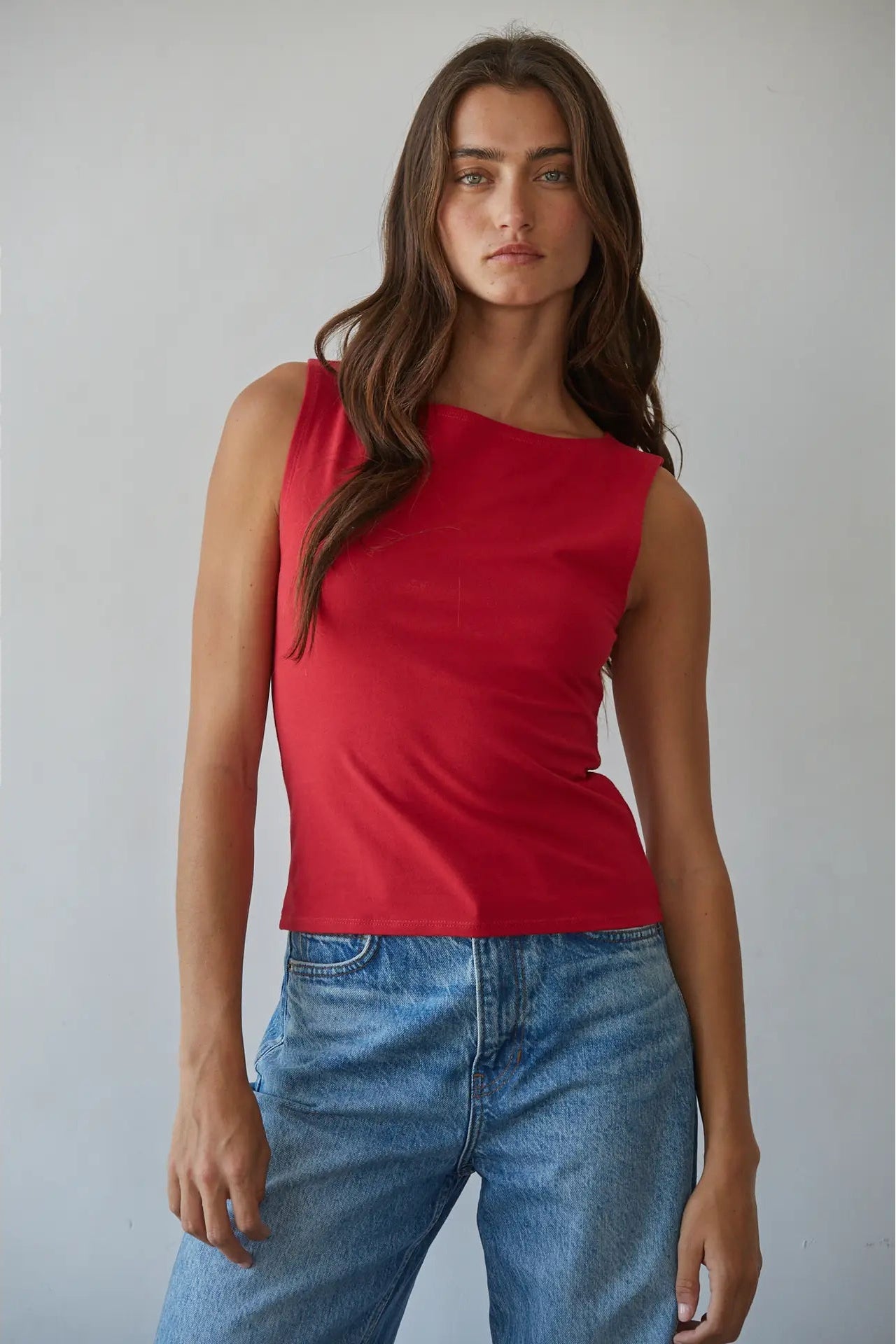 A woman modeling a sleeveless red top with an asymmetrical neckline and blue jeans against a plain light gray studio background.