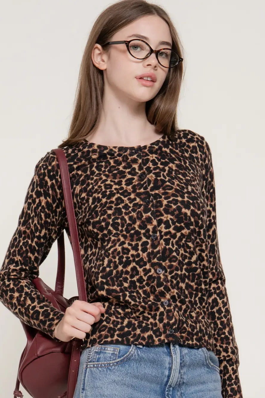 A woman wearing a brown and black leopard print long-sleeve knit top, paired with blue jeans and a burgundy shoulder bag, against a plain off-white background.