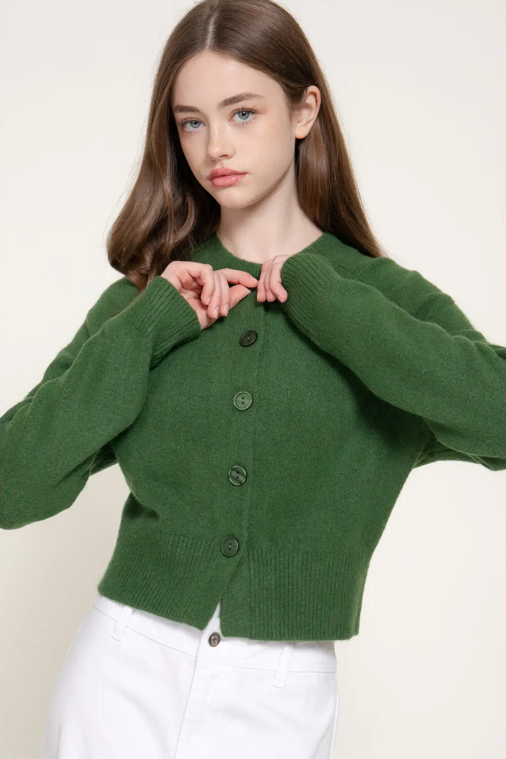 A woman wearing a cropped green knit cardigan with dark buttons and white pants, posed against a plain light beige studio background.