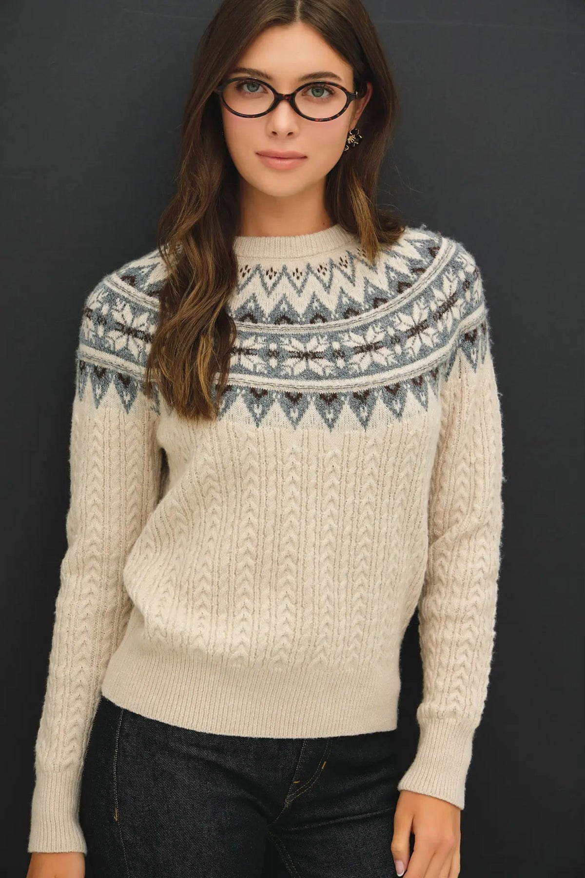 A woman wearing a cream-colored cable-knit sweater with a gray and blue Fair Isle yoke pattern, paired with dark jeans, set against a dark gray background.