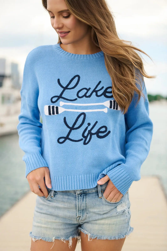 Woman wearing a blue 'Lake Life' sweater by a waterfront.