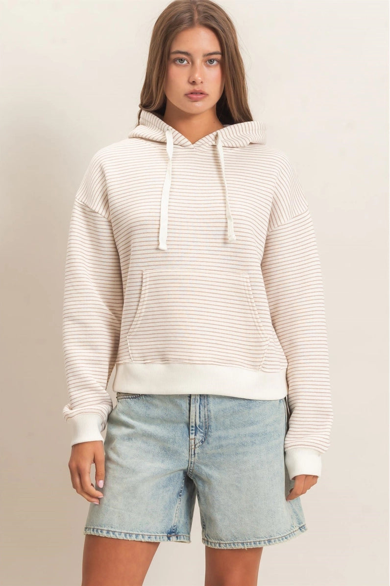 A woman wearing a cream and white horizontally striped pullover hoodie with white drawstrings and a front kangaroo pocket, paired with light-wash blue jeans, set against a plain light beige studio background.