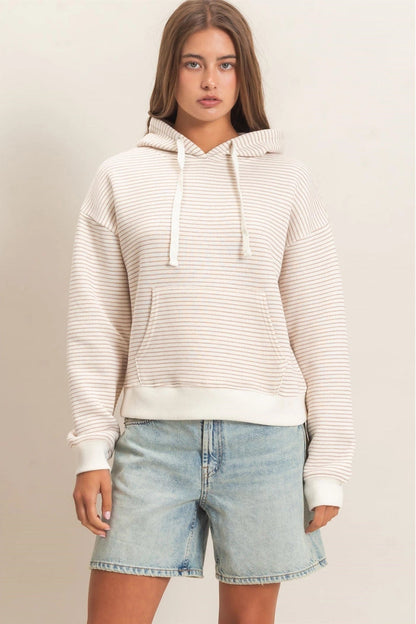 A woman wearing a cream and white horizontally striped pullover hoodie with white drawstrings and a front kangaroo pocket, paired with light-wash blue jeans, set against a plain light beige studio background.
