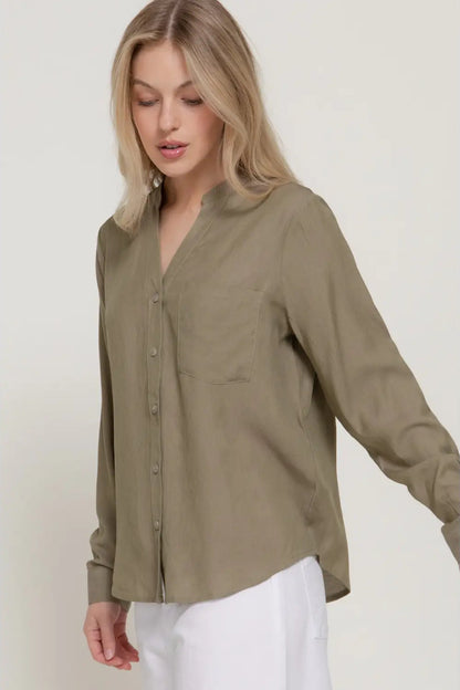 A woman wearing a long-sleeve button-front blouse in a muted olive khaki color, paired with white pants, against a plain light beige studio background.