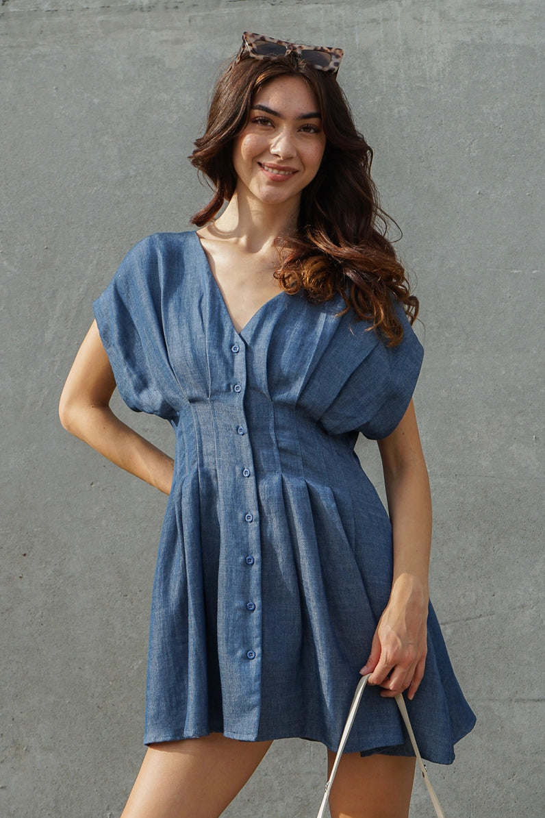 A woman wearing a blue denim-like mini dress with puff sleeves, a V-neck, and a button front, standing against a textured gray wall.