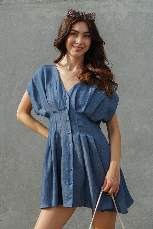 A woman wearing a blue denim-like mini dress with puff sleeves, a V-neck, and a button front, standing against a textured gray wall.