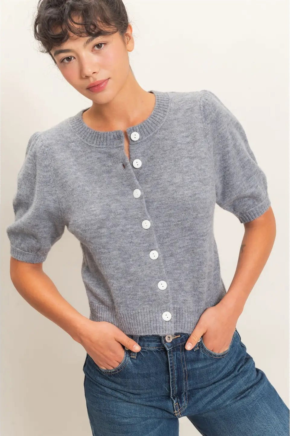 A woman wearing a gray short-sleeve knit cardigan with white buttons down the front, paired with blue jeans, standing against a plain light beige background.