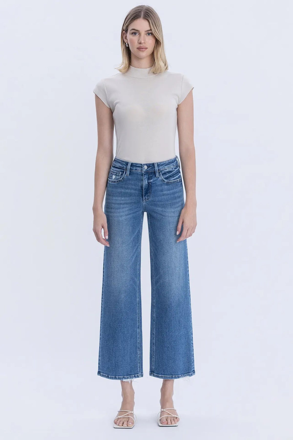 A woman modeling a beige short-sleeve mock neck top and blue wide-leg cropped jeans against a plain white studio background.