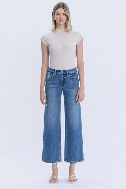 A woman modeling a beige short-sleeve mock neck top and blue wide-leg cropped jeans against a plain white studio background.