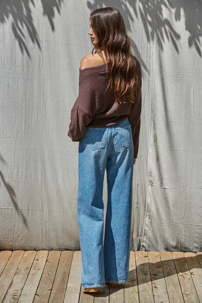 A woman stands with her back to the camera wearing a brown off-the-shoulder long-sleeve top and light blue wide-leg jeans, standing on a wooden deck in front of a gray fabric backdrop.