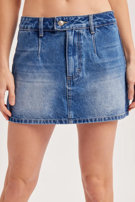 A close-up of a model wearing a blue denim mini skirt with a faded wash, button closure, and belt loops against a light studio background.