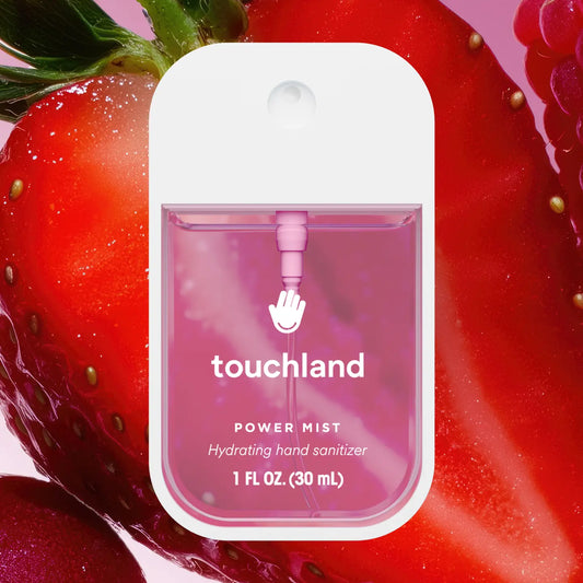 A Touchland Power Mist hydrating hand sanitizer dispenser bottle, featuring a white cap and a pink translucent body, set against a vibrant, close-up background of fresh strawberries.