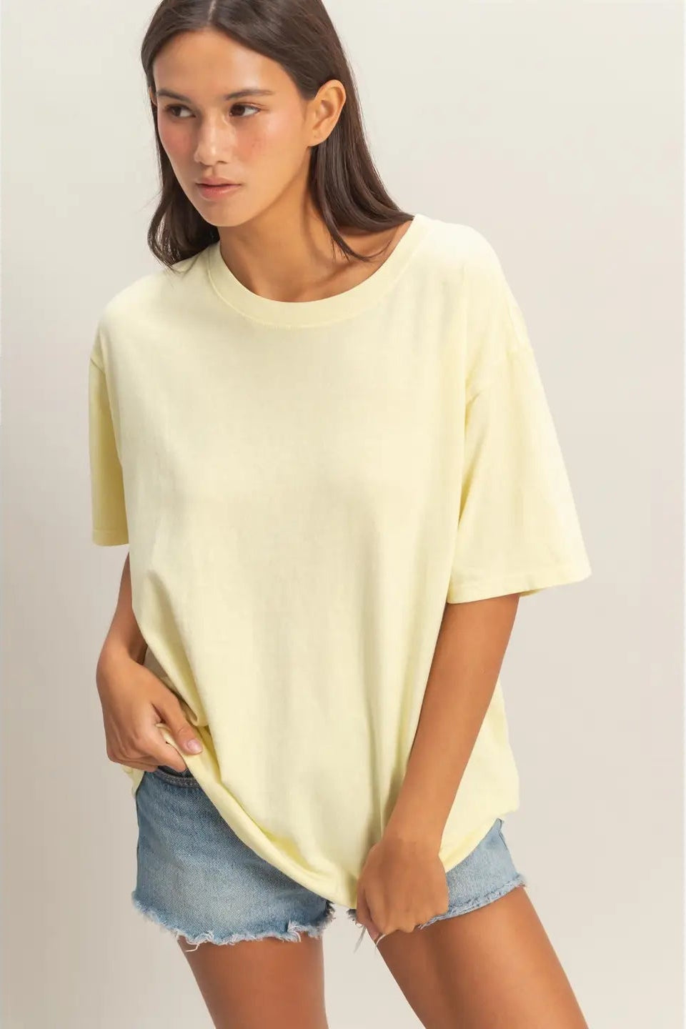 Woman wearing a light yellow oversized t-shirt and denim shorts on a plain background