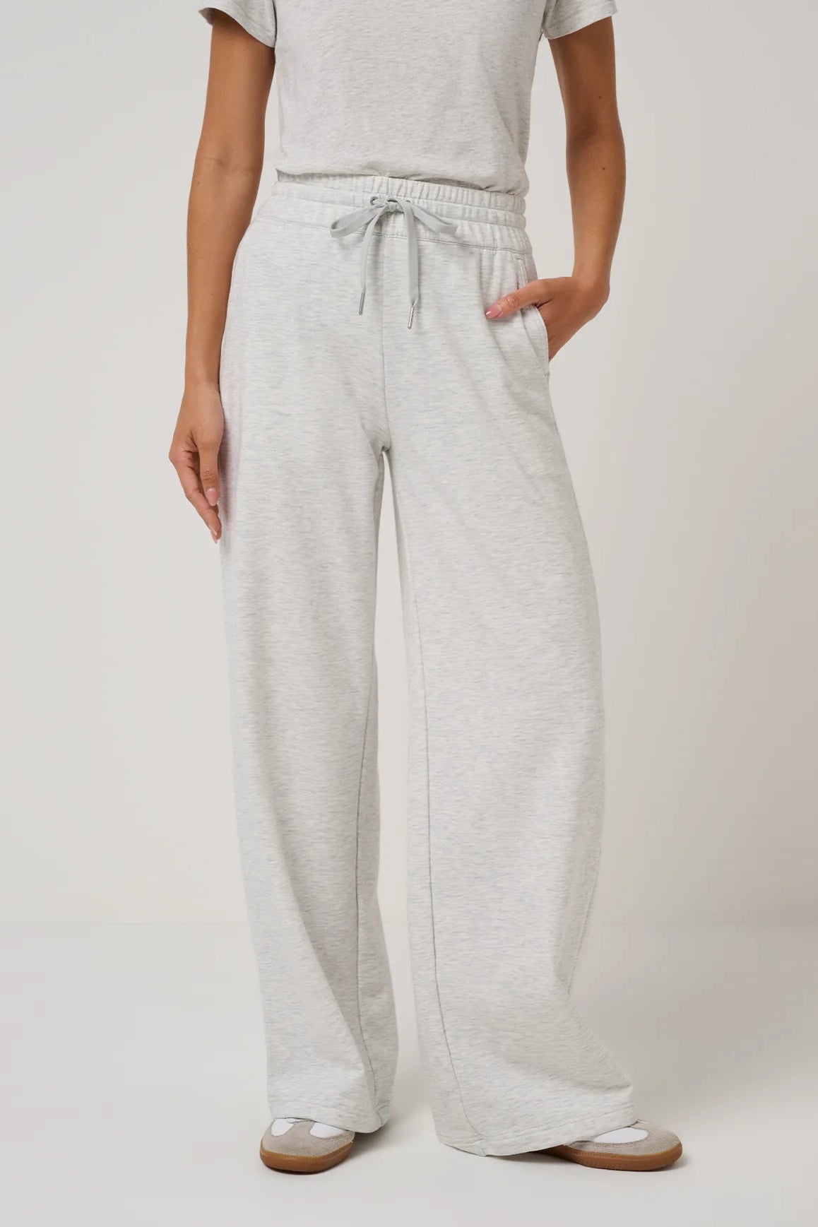 A person wearing light heather gray wide-leg sweatpants with an elastic drawstring waist, paired with a matching light gray top and beige sandals, shown against a plain light gray studio background.