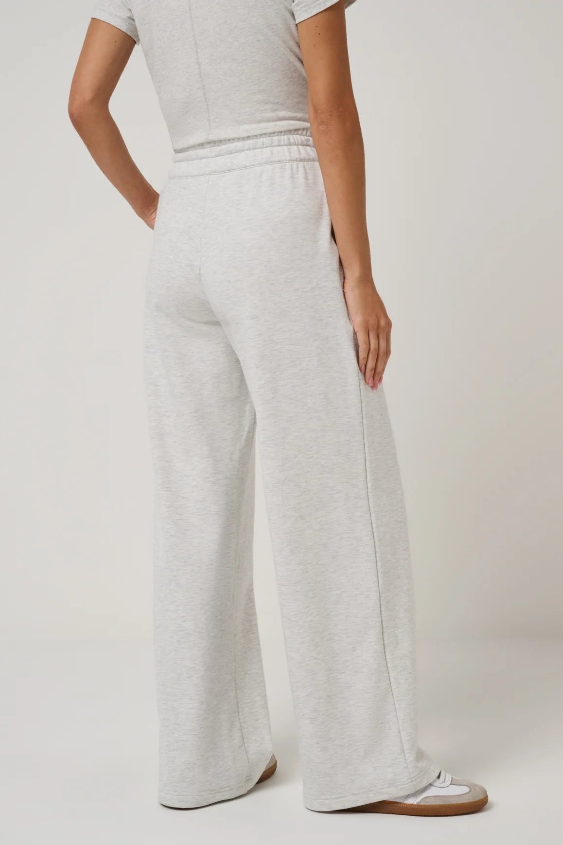 A person shown from the back wearing light heather gray wide-leg sweatpants with an elastic waistband, paired with a matching light gray top and white sneakers, against a plain light gray studio background.