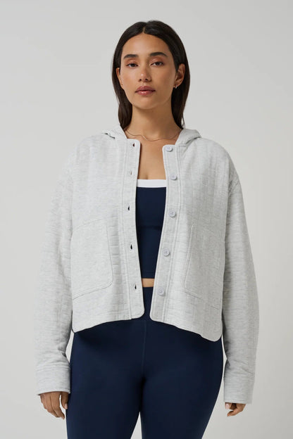 A woman modeling a light gray heather cropped button-up hooded jacket over a navy blue sports bra and navy leggings, shot against a solid light gray studio background.