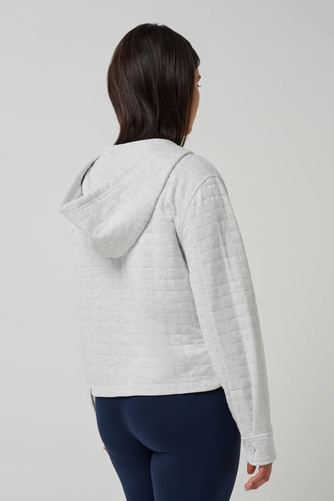 A woman shown from the back wearing a light gray heather cropped hooded jacket with a quilted texture, paired with navy blue leggings, against a plain light gray studio background.