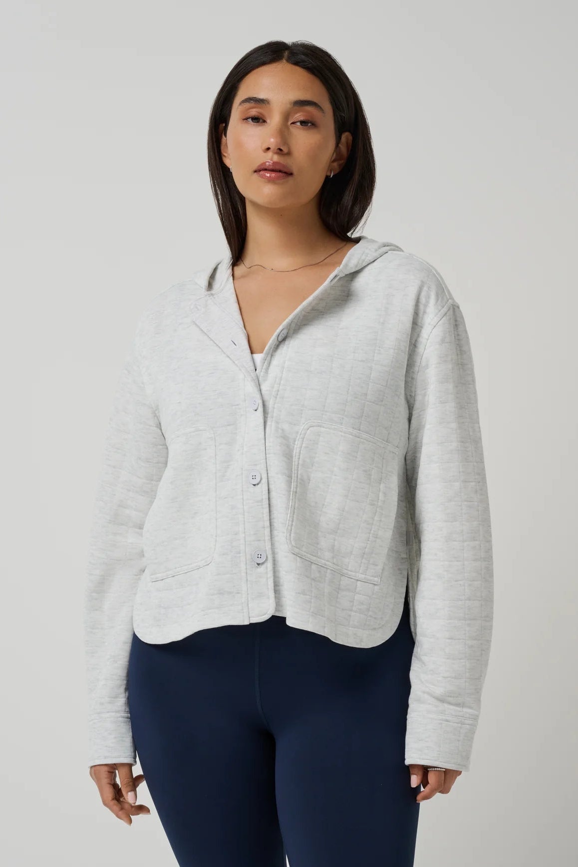 A woman modeling a light heather gray cropped button-up cardigan with a hood and front pockets, paired with dark navy leggings, against a plain light gray studio background.