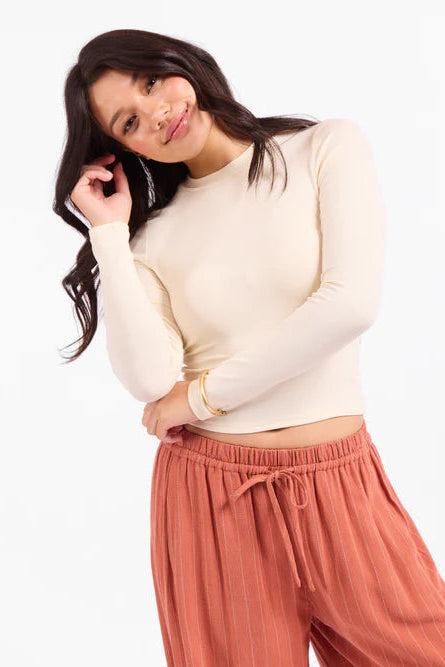 A woman posing in a studio setting, wearing a fitted, long-sleeve cream mock-neck crop top and dusty rose wide-leg drawstring pants.