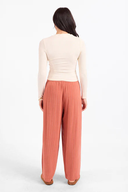 A woman viewed from the back wearing a fitted cream long-sleeve top and wide-leg dusty rose pants against a plain white studio background.