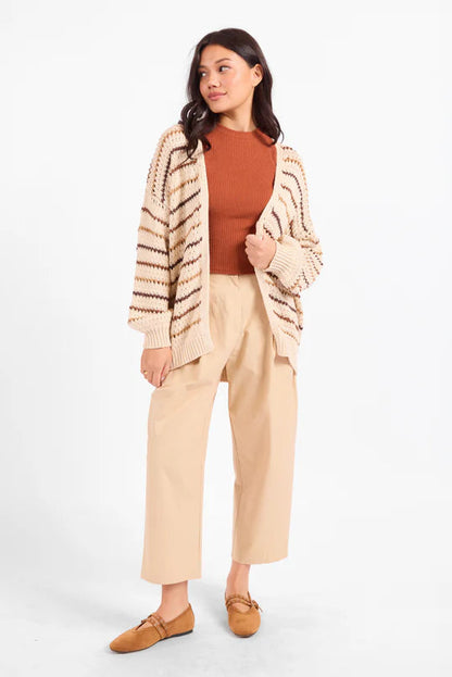 A woman modeling a beige and brown striped knit cardigan over a rust-orange ribbed top, paired with beige cropped pants and tan loafers, against a plain white studio background.