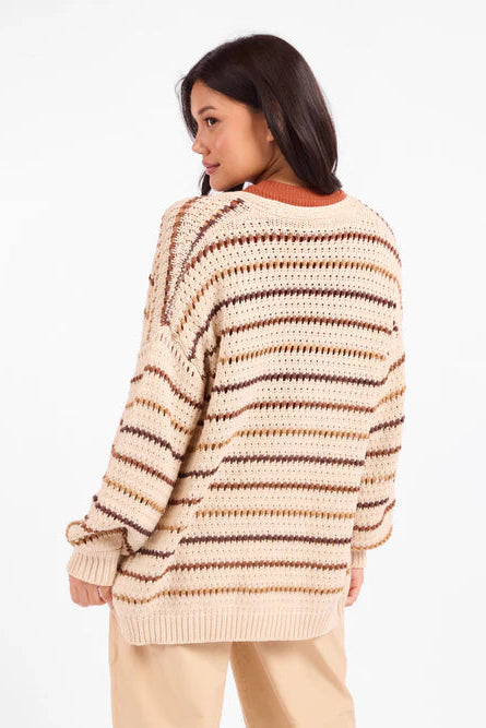 A woman wearing a chunky knit sweater with horizontal beige, brown, and dark brown stripes, shown from a three-quarters back view against a plain white background.