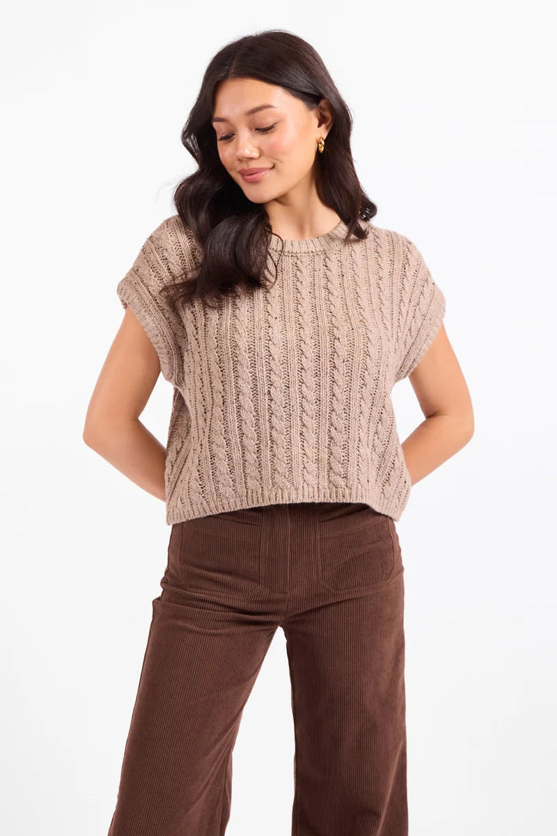 A woman wearing a beige cable-knit short-sleeve cropped sweater and brown corduroy pants, standing against a plain white studio background.