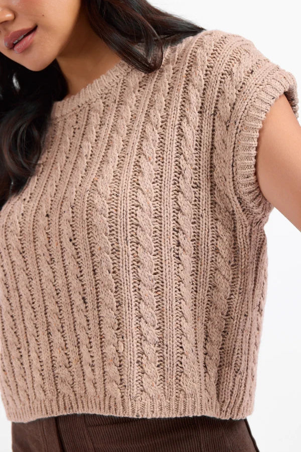 A close-up of a woman wearing a cropped, short-sleeve cable-knit sweater in a warm taupe-beige color, paired with brown corduroy pants against a plain white background.