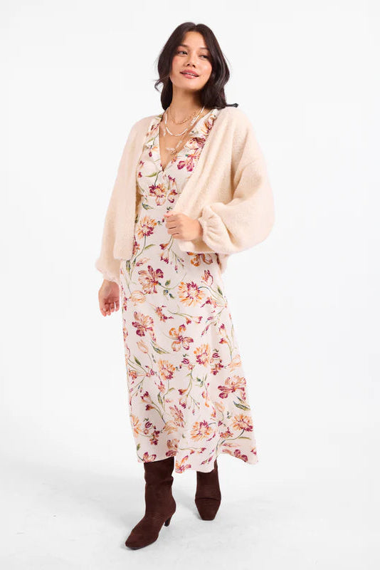 A woman modeling a long-sleeve cream cardigan over a floral midi dress with brown pointed-toe boots, photographed against a plain white studio background.