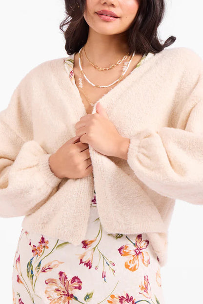 A close-up of a woman wearing a fuzzy cream-colored cropped cardigan with voluminous sleeves, layered over a floral dress.