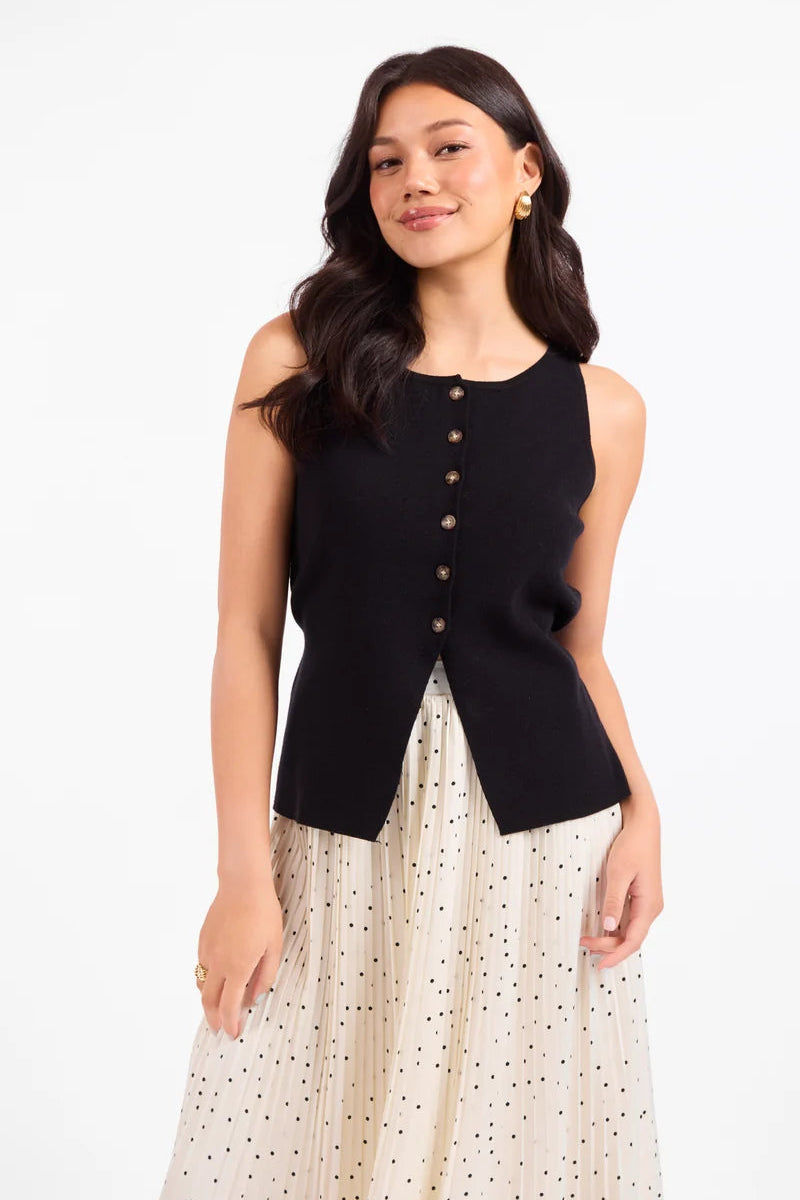 A woman modeling a sleeveless black knit vest with a button-down front, paired with a cream polka-dot skirt, against a plain white studio background.