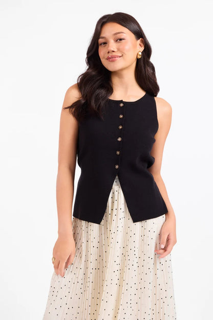 A woman modeling a sleeveless black knit vest with a button-down front, paired with a cream polka-dot skirt, against a plain white studio background.
