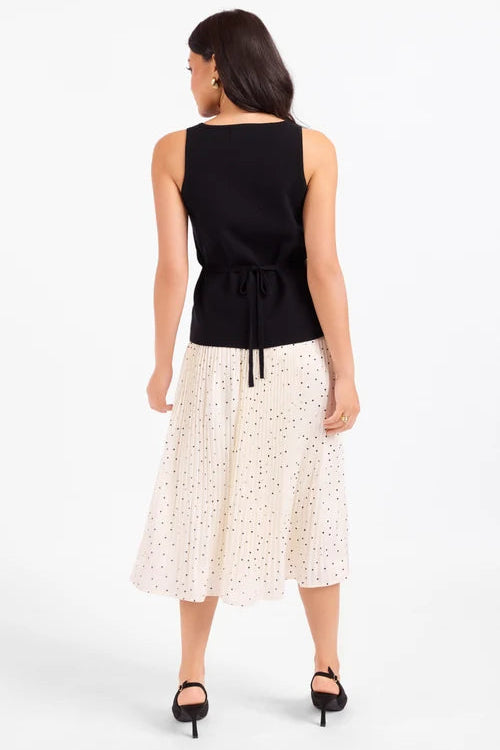 A woman shown from the back wearing a sleeveless black top with a tie waist and a cream midi skirt with small black polka dots, paired with black heels, against a plain white studio background.