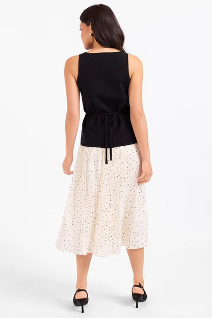 A woman shown from the back wearing a sleeveless black top with a tie waist and a cream midi skirt with small black polka dots, paired with black heels, against a plain white studio background.