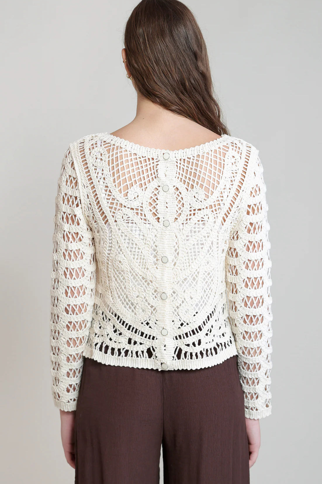 A woman viewed from the back wearing a cream crochet lace long-sleeve top with a scoop neckline and buttoned back, paired with dark brown pants, against a light gray studio background.