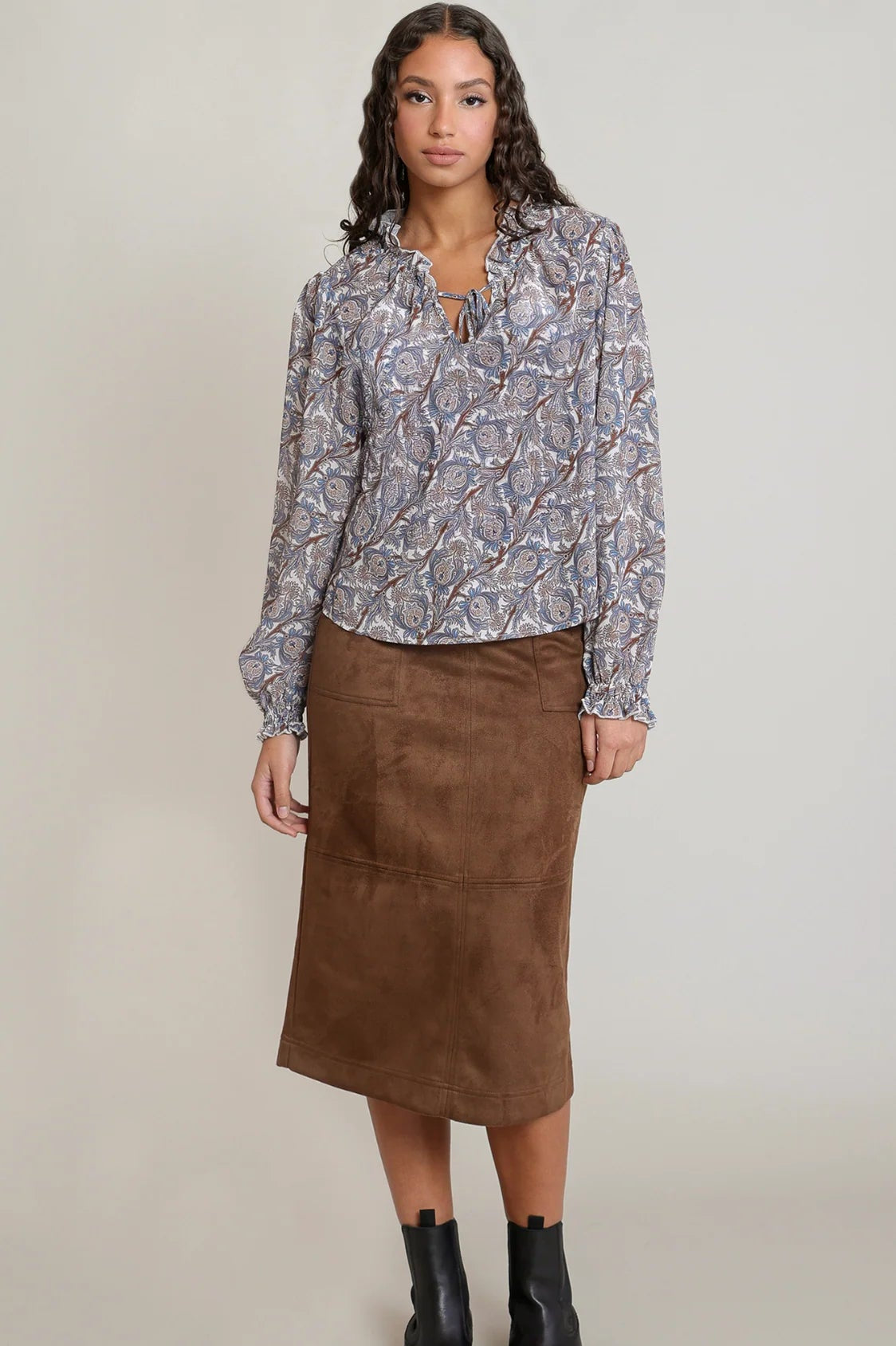 A woman modeling a long-sleeve button-up blouse with a blue, gray, and white abstract pattern, paired with a brown midi skirt and black ankle boots, against a plain light gray studio background.