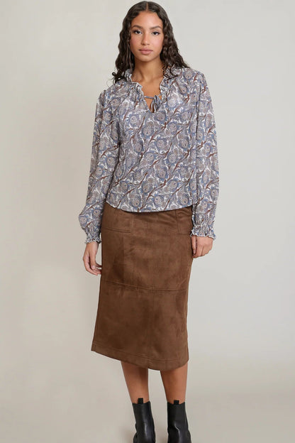 A woman modeling a long-sleeve button-up blouse with a blue, gray, and white abstract pattern, paired with a brown midi skirt and black ankle boots, against a plain light gray studio background.