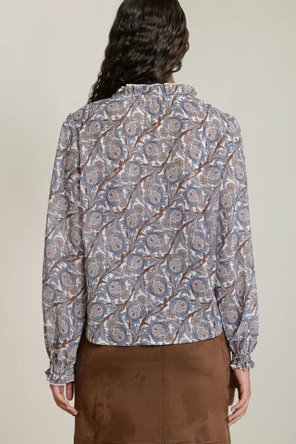 A woman shown from the back wearing a long-sleeve button-up blouse with an intricate paisley pattern in blue, beige, and brown, paired with a brown skirt, against a neutral studio background.