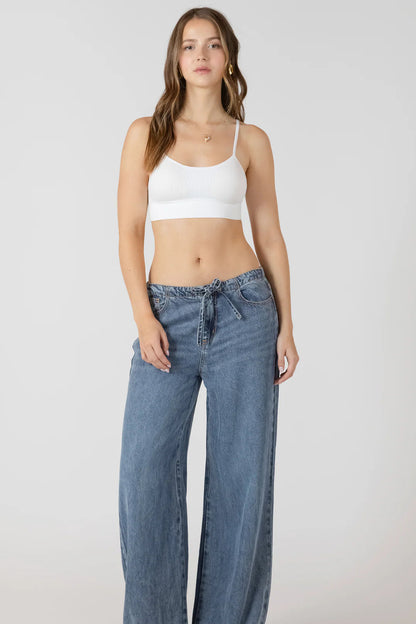 Woman wearing a white tank top and blue wide-leg jeans on a plain background