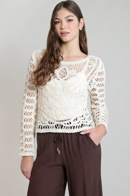 A woman modeling a cream-colored, long-sleeve crochet knit top paired with brown drawstring pants against a light gray studio background.