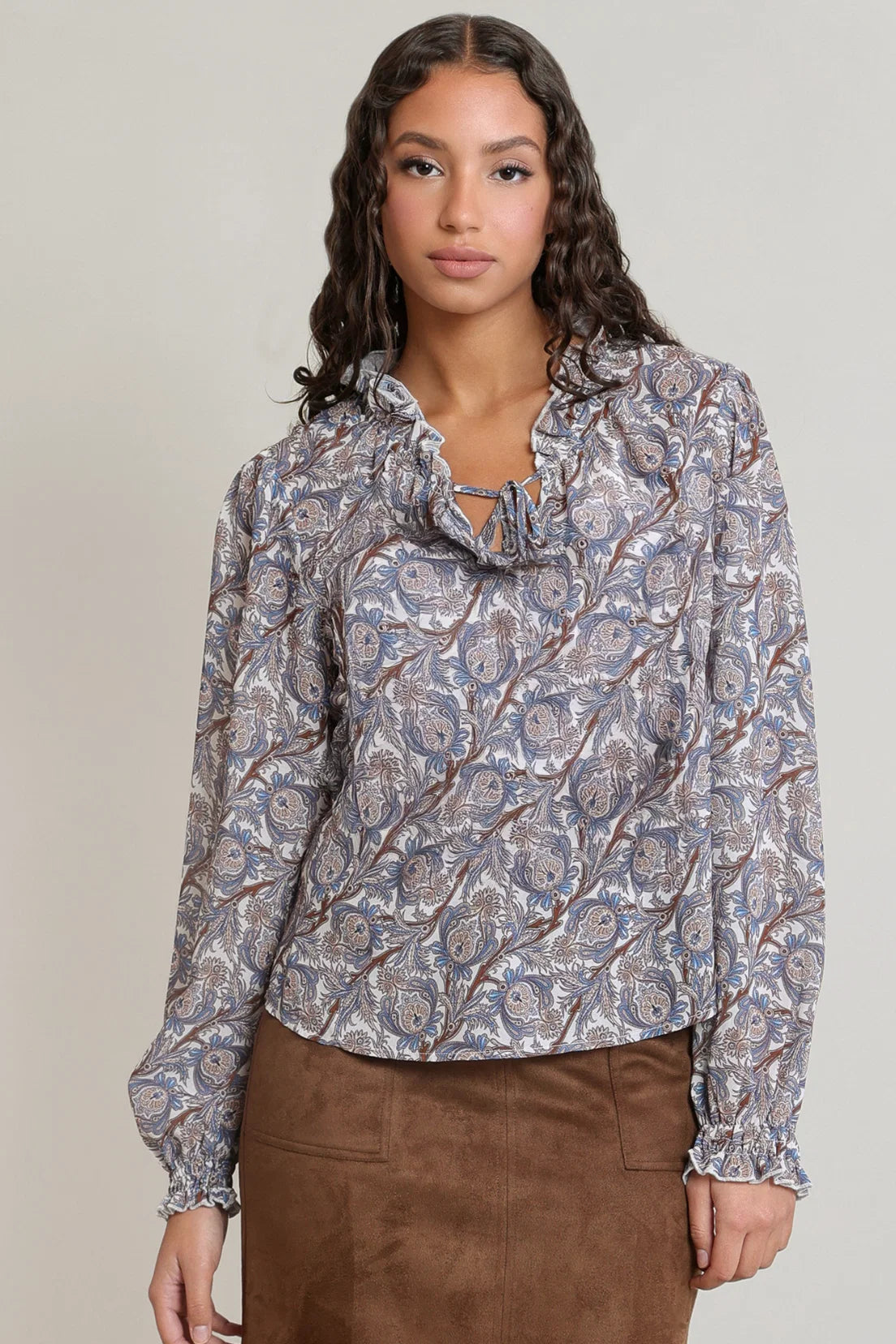 A woman wearing a long-sleeve blouse with a blue, gray, and brown paisley pattern and a ruffled tie-neck detail, paired with a brown skirt, against a plain light gray background.