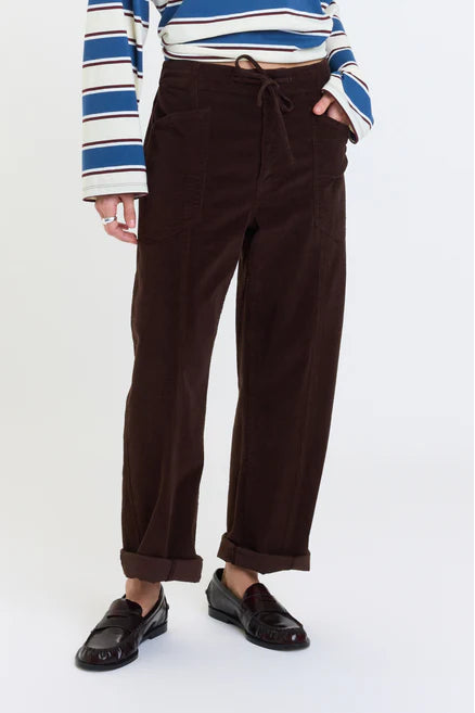 A person wearing dark brown wide-leg corduroy trousers with a drawstring waist, paired with a blue and white striped long-sleeve top and black loafers, shown from the waist down on a light gray studio background.