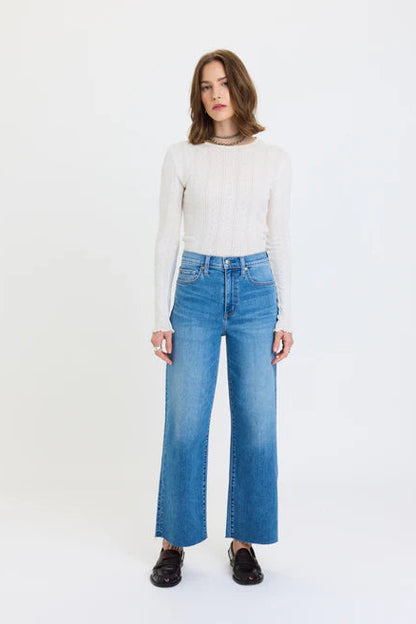 A woman modeling a cream ribbed long-sleeve top and high-waisted, wide-leg blue jeans against a plain white studio background.